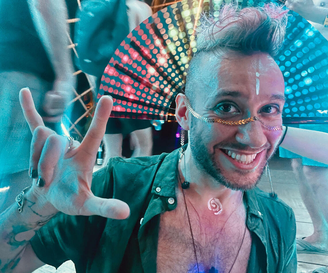 Photo of a man resting while on a hike in the mountainside, a blue lake and mountains in the background, sunset hour coloring clouds in orange and pink hues.