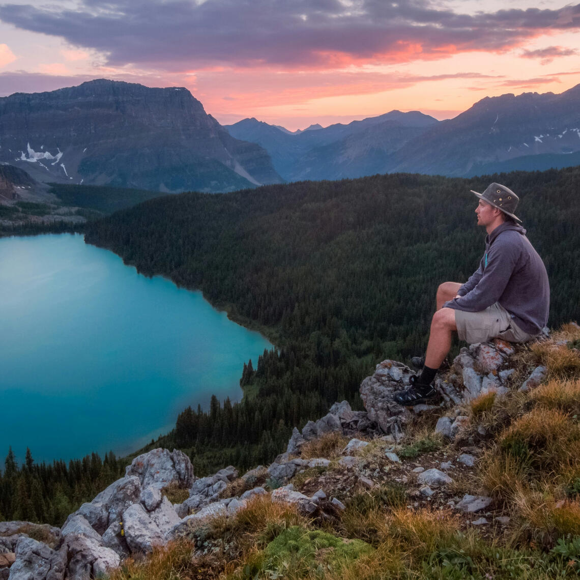 Photo of a man resting while on a hike in the mountainside, a blue lake and mountains in the background, sunset hour coloring clouds in orange and pink hues.